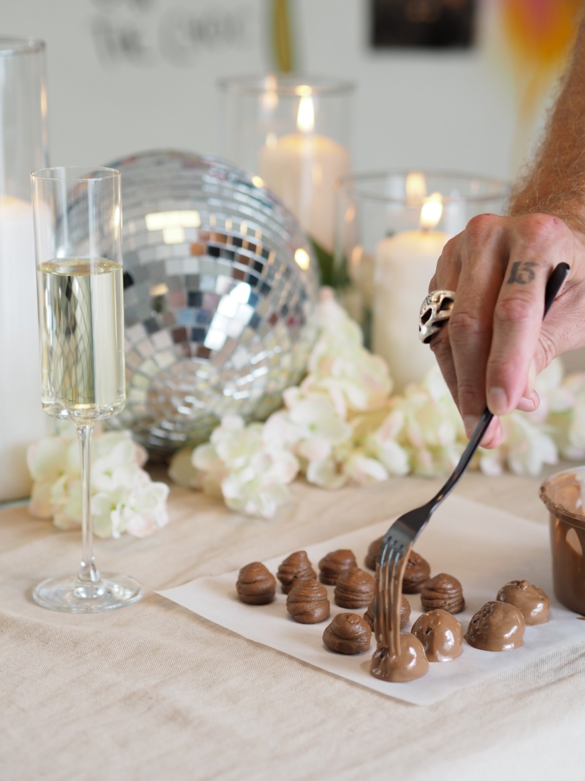 A table decorate with flowers, a disco ball and candles with a sheet of chocolate truffles on it and a person's hand holding a fork